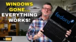 Graeme from tektoc holding an Asus BR1100 2-in-1 touch screen laptop running Fedora Linux, with the text "Windows Gone… Everything Works!"